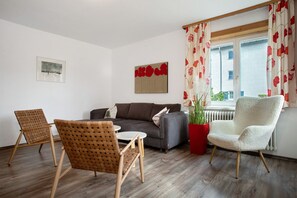 Ein Wohnzimmer mit einer Couch, zwei Sessel, einem kleinen Tisch, einer Zimmerpflanze und einem Fenster mit Vorhängen.