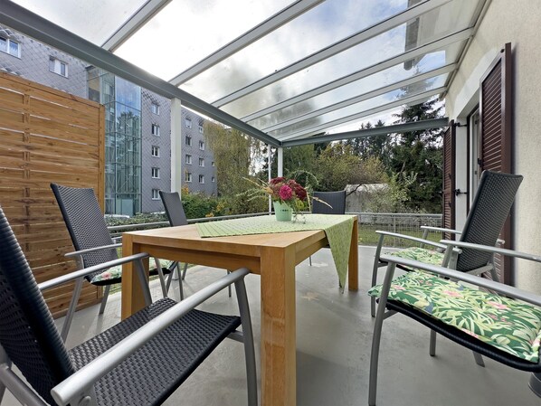 Eine überdachte Terrasse mit Tisch und Stühlen, ein Holzzaun und ein Gebäude im Hintergrund.