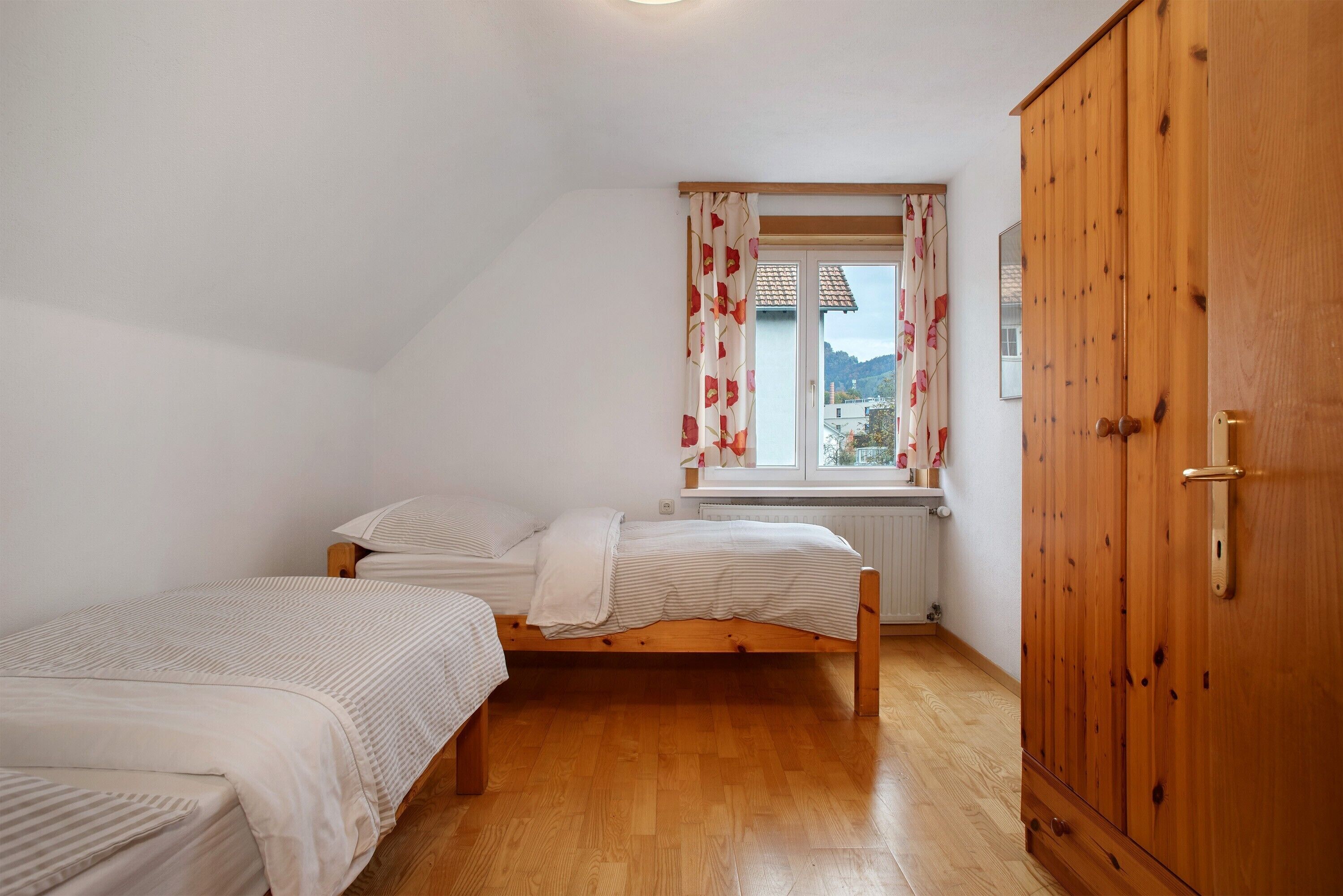 Ein Schlafzimmer mit zwei Einzelbetten, einem Holzschrank, einem Fenster mit Vorhängen und einer Heizung.