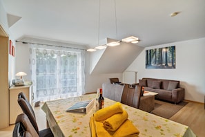 A modern living room with a dining area, a sofa, a table with a book and a bottle, and a forest-themed painting on the wall.