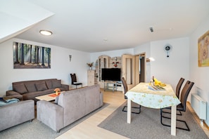 A modern living room with a sofa, a dining table with chairs, a flat-screen TV, and a kitchen area with cabinets and a refrigerator.