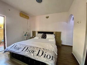 A bedroom with a large bed, a bedside table, a fan, and a wall-mounted heater.