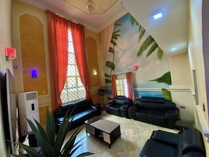 A living room with a sofa, two armchairs, a coffee table, and a fan. There is a large window with curtains, a wall-mounted air conditioner, and a ceiling fan.