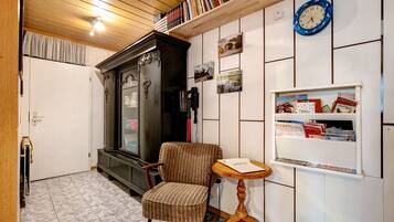 A room with a wooden ceiling, a radiator, a striped armchair, a small wooden table, a wall clock, and a bookshelf with books and magazines.