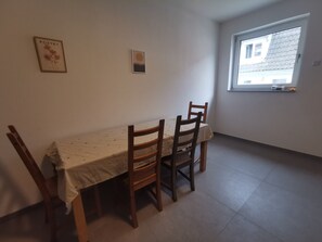 Un coin repas avec une table et des chaises, un tableau encadré au mur et une fenêtre donnant sur des immeubles.