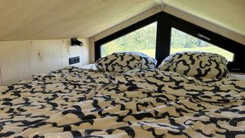 Une chambre avec un plafond en pente, un lit recouvert d’une couvre-lit à motifs, deux fenêtres et un luminaire fixé au mur.