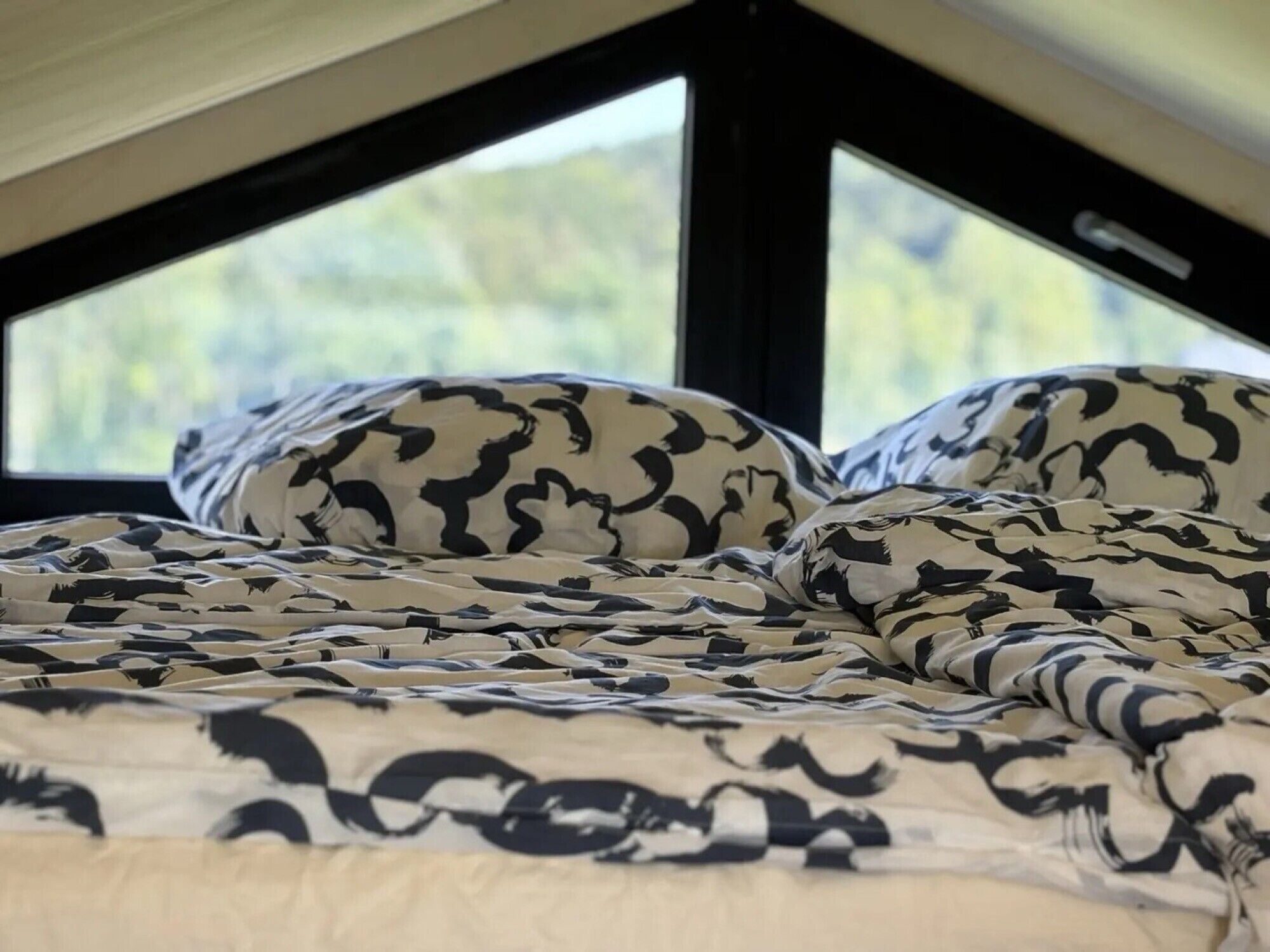 Ein Bett mit gemusterten Betttextilien, ein Fenster mit Blick ins Grüne und ein schwarzer Rahmen.