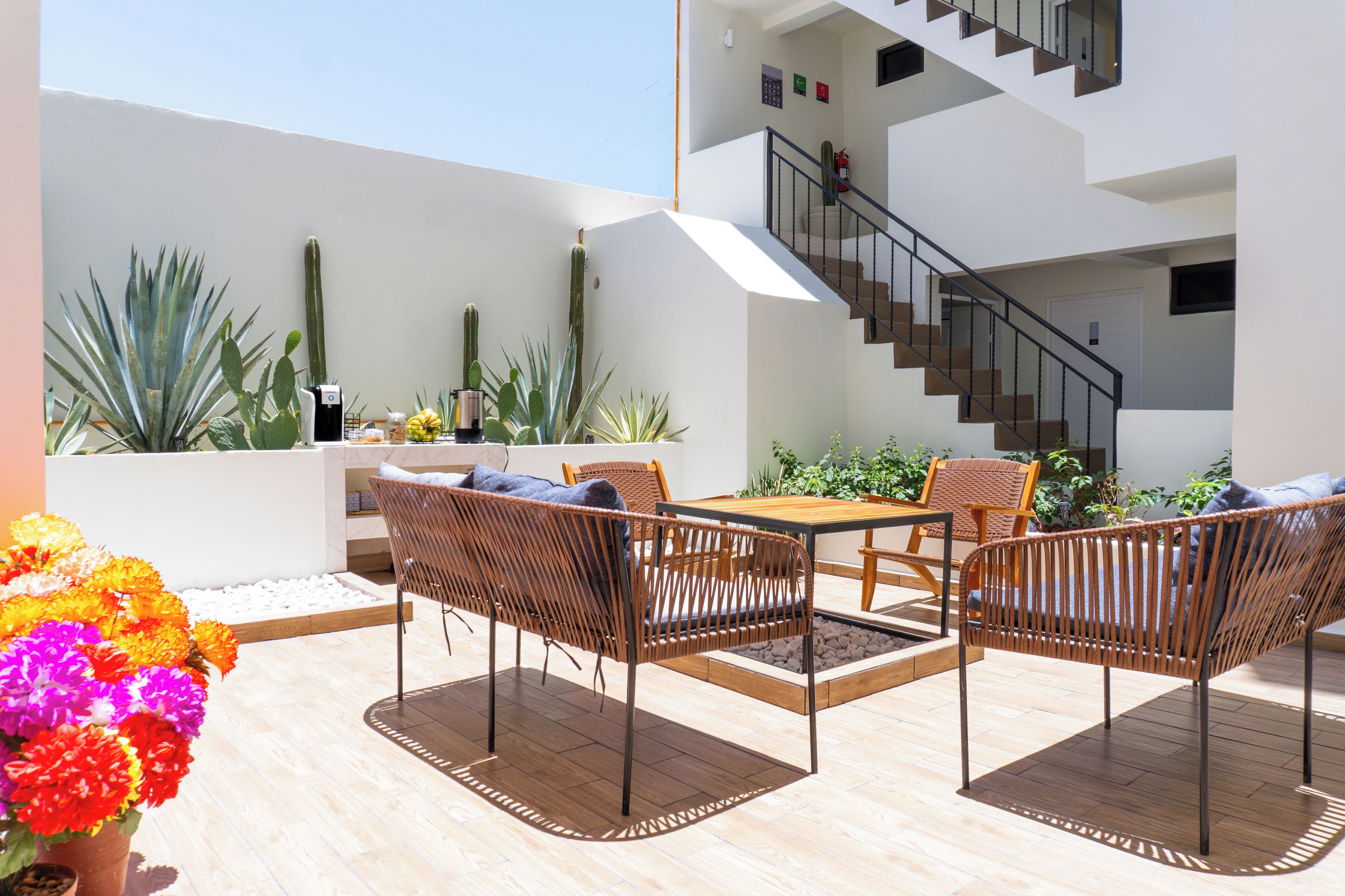 Een buitenpatio met rieten meubels, een houten tafel en diverse planten, waaronder cactussen.