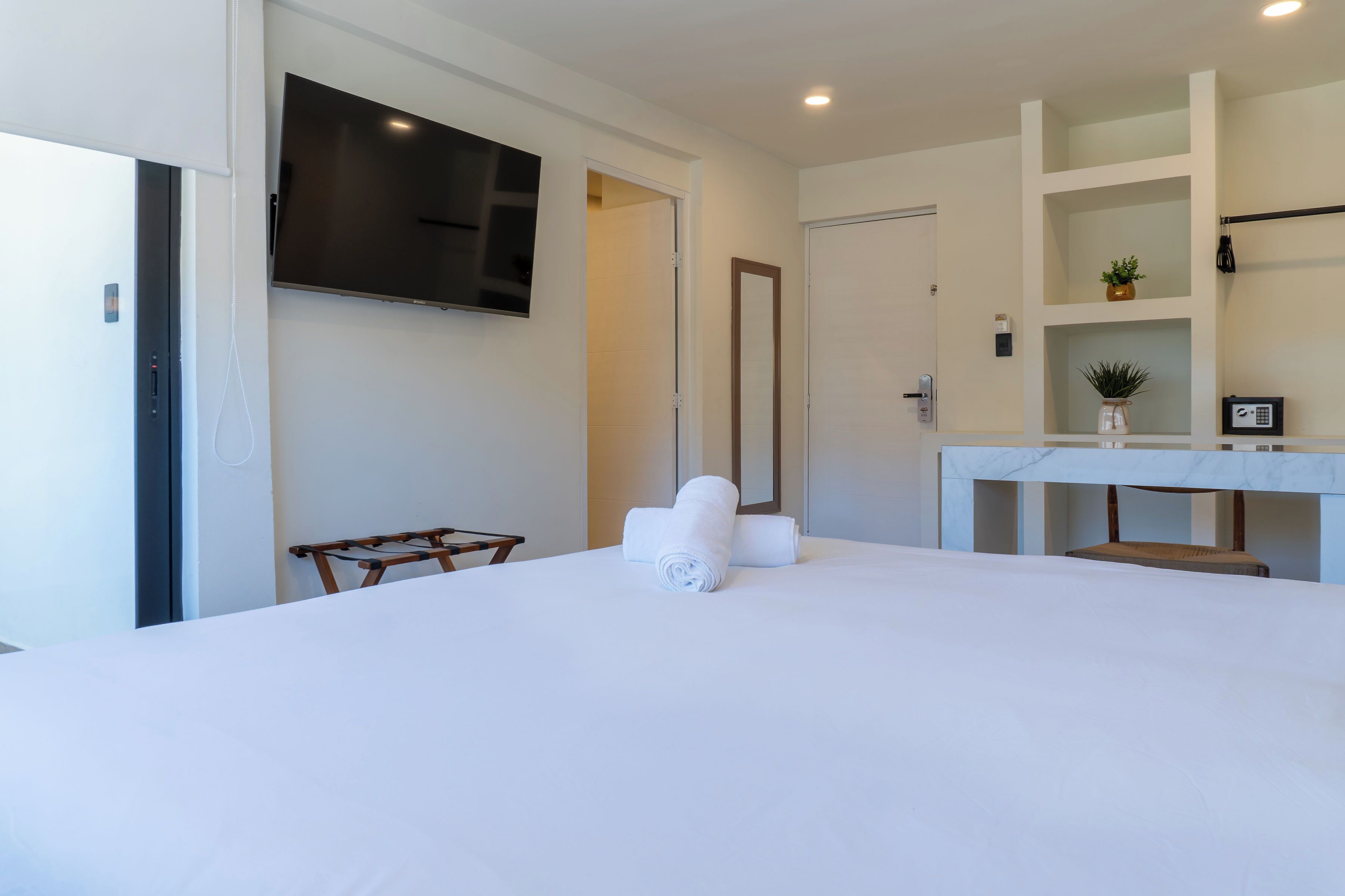 A hotel room with a large bed, a TV mounted on the wall, a desk with a chair, and a small plant on the shelf.