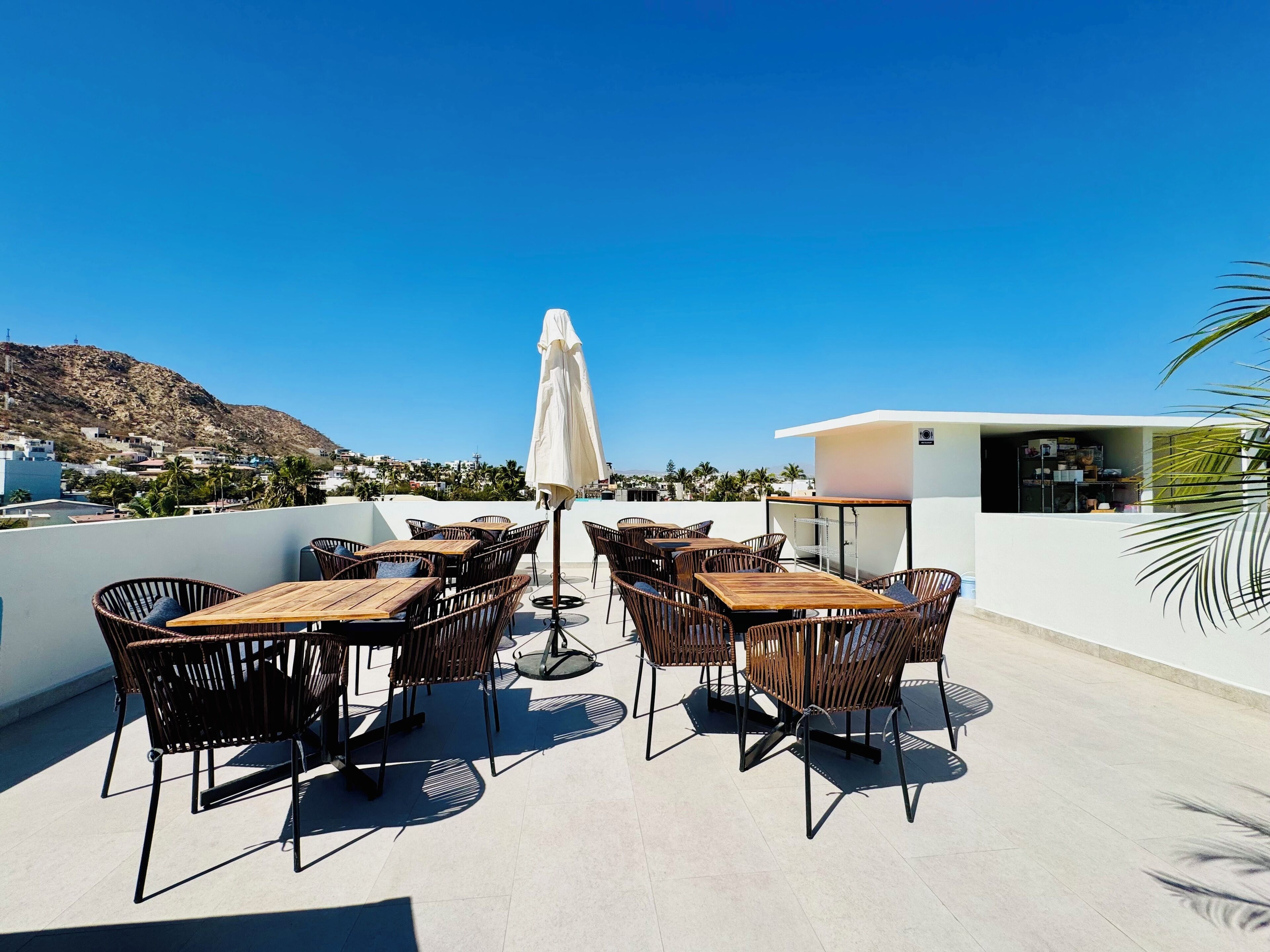A rooftop terrace with wooden tables and wicker chairs, a white umbrella, and a view of a hillside town.