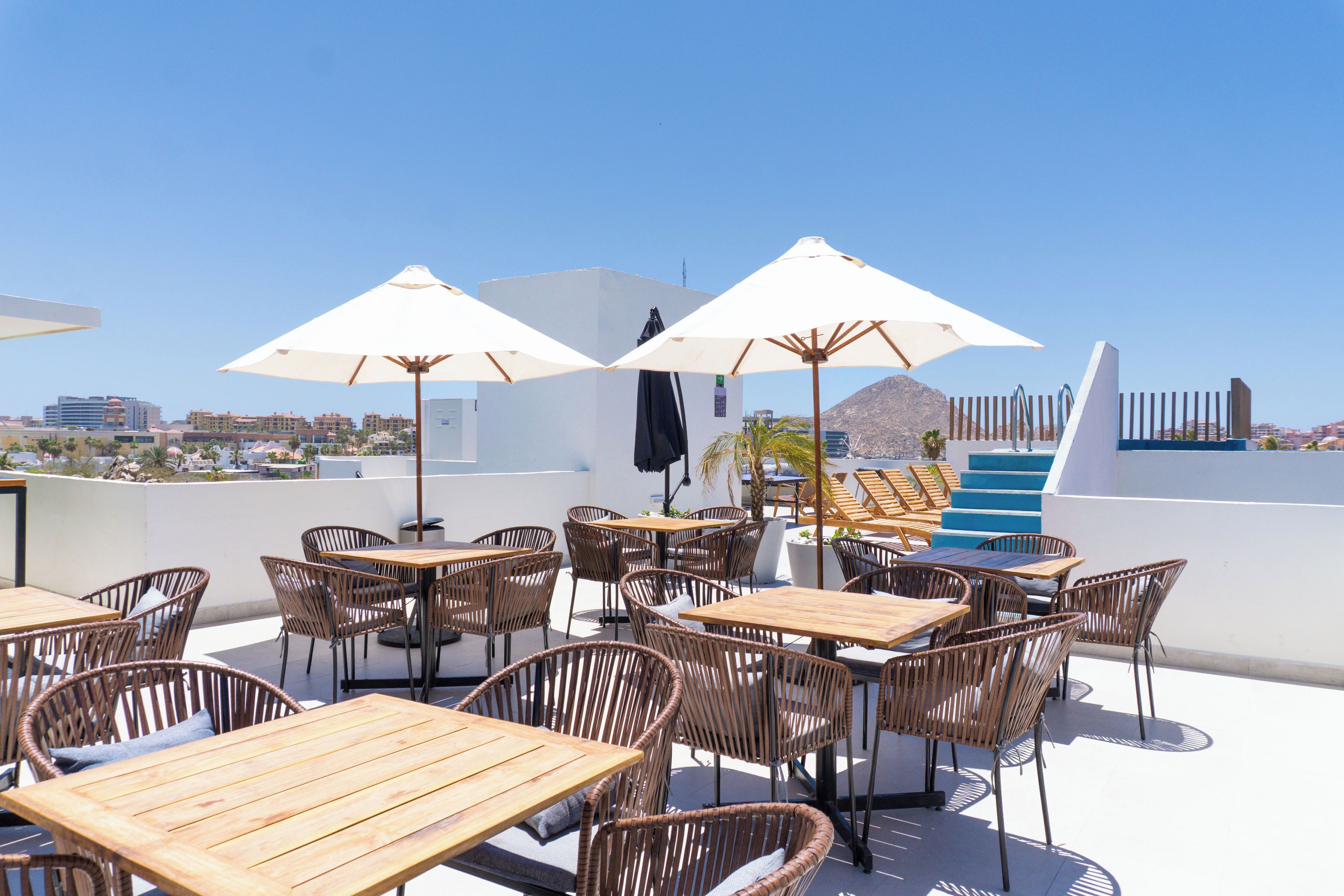 An outdoor seating area with wooden tables and wicker chairs, white umbrellas, and a view of a cityscape in the background.
