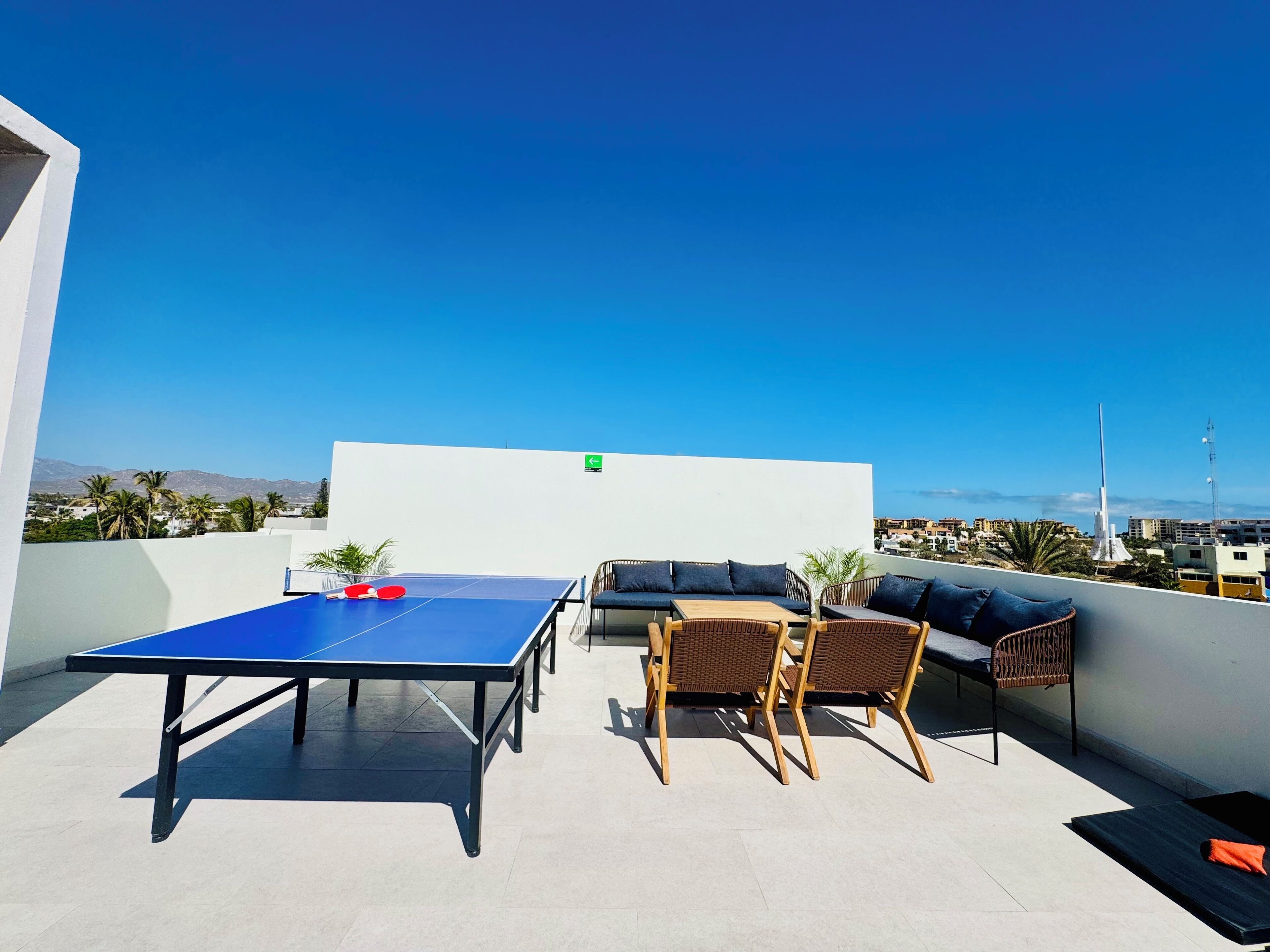 A rooftop terrace with a ping pong table, wicker furniture, and a view of the city and mountains.