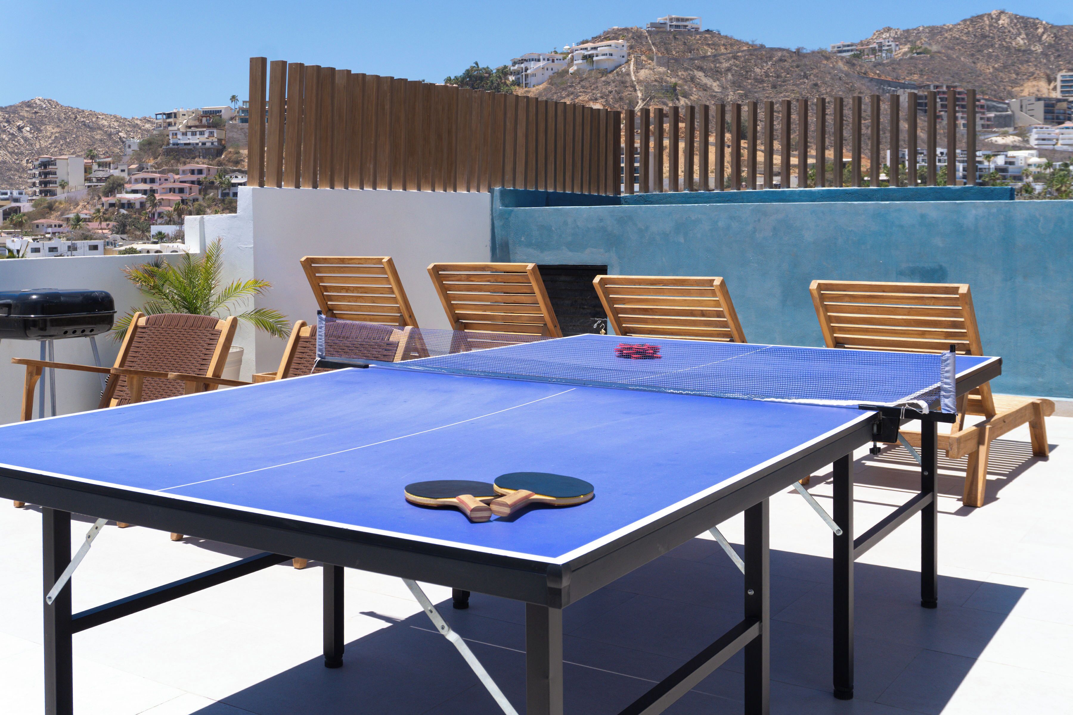 A ping pong table with two paddles on a rooftop terrace with wooden chairs and a view of a hillside town.