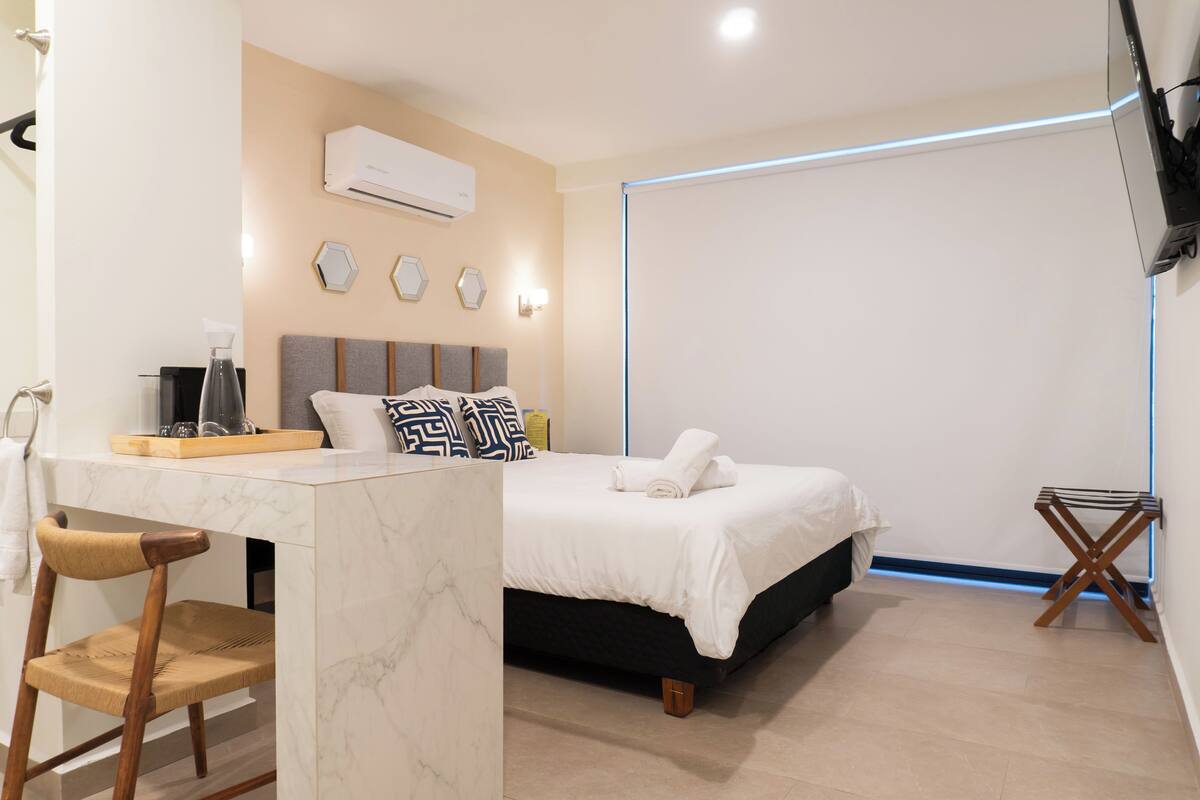 A modern hotel room with a bed, a desk with a chair, a small table, and a TV mounted on the wall.