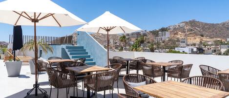 An outdoor patio with wooden tables and wicker chairs, large umbrellas, and a staircase leading to an upper level.