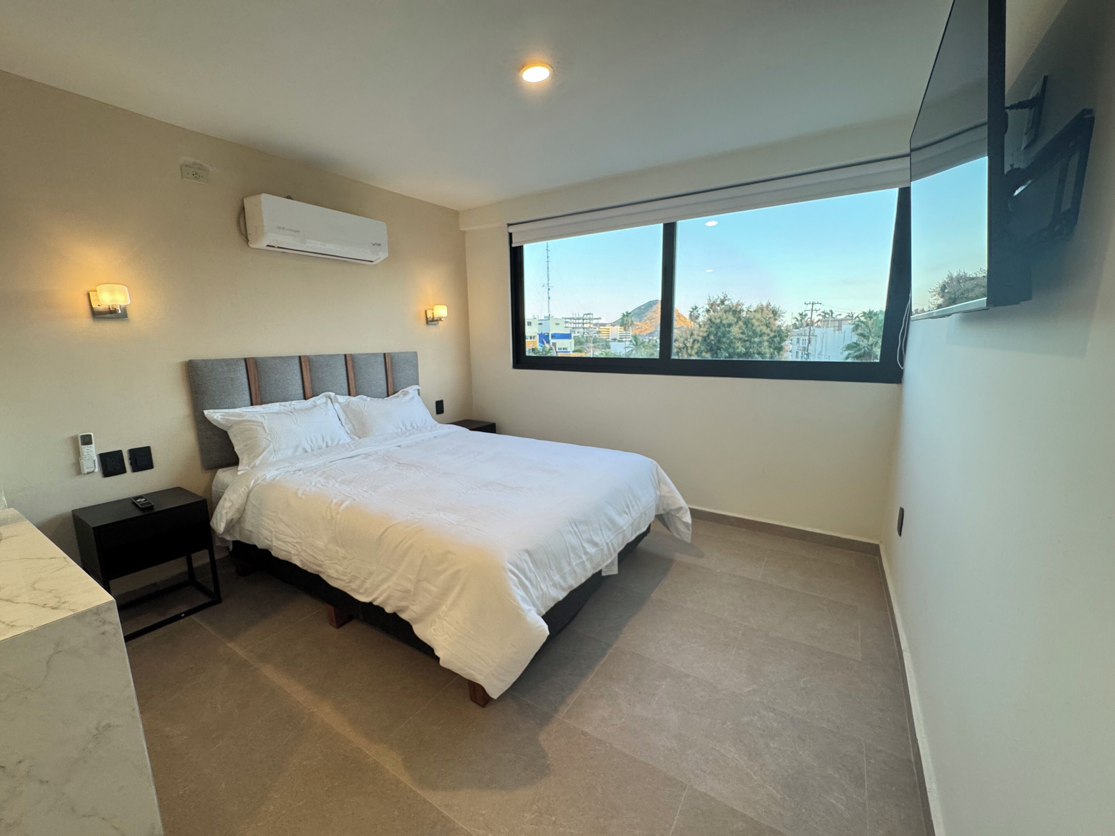 A modern bedroom with a large bed, a nightstand, a wall-mounted air conditioner, and a window with a city view.