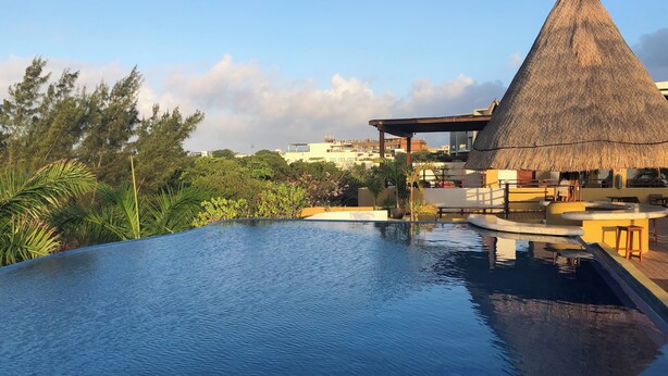 A swimming pool with a wooden deck, a thatched-roof structure, and surrounding greenery. - 2 BR with infinity adult pool + family pool, ocean view, & best street in Playa! (Playa del Carmen)