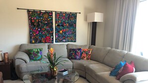 A living room with a beige sofa, colorful pillows, a glass coffee table with a plant, and two vibrant wall hangings. - 2 BR with infinity adult pool + family pool, ocean view, & best street in Playa! (Playa del Carmen)