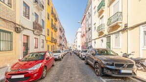 A cobblestone street lined with colorful buildings, parked cars, and a clear blue sky. - GuestReady - One prime stay in Lisbon (Lisbon)
