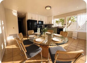 A dining area with a glass table surrounded by chairs, a kitchen in the background, and a window with curtains.