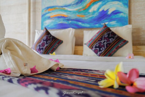 A bed with patterned pillows, a colorful woven mat, and a painting of a wave on the wall. - Java Wood Villa (Gili Trawangan)