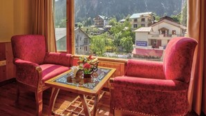A room with two red upholstered chairs, a wooden table with a vase of flowers, and a large window offering a view of a mountain and a village. - Hotel Swastik Grand (Manali)