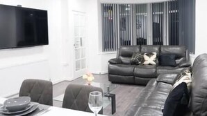 Una sala de estar moderna con un sofá de cuero negro, un televisor de pantalla plana montado en la pared y un comedor con una mesa de vidrio.
