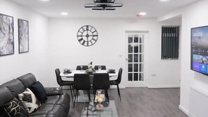 Una sala de estar moderna con un sofá de cuero negro, una mesa de centro de vidrio y un comedor con una mesa blanca y sillas negras. Hay un reloj de pared y un televisor de pantalla plana montado en la pared.