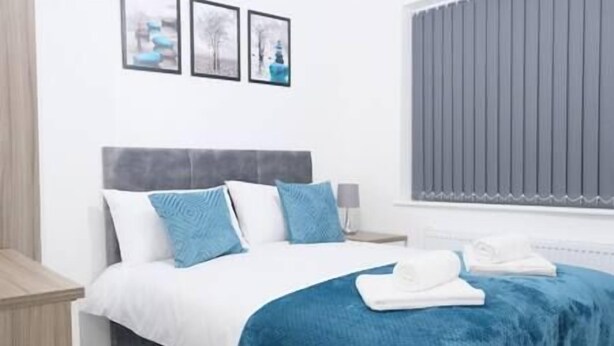 Una cama bien hecha con ropa de cama azul y blanca, un cabecero gris y una ventana con persianas verticales.