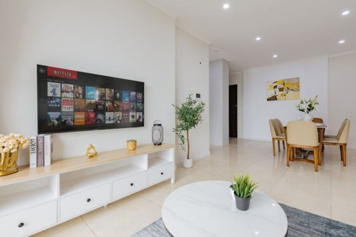 Un salon moderne doté d’un téléviseur à écran plat fixé au mur, d’une console en bois agrémentée d’objets décoratifs, d’une table basse ronde avec une plante, et d’un coin repas avec une table et des chaises.