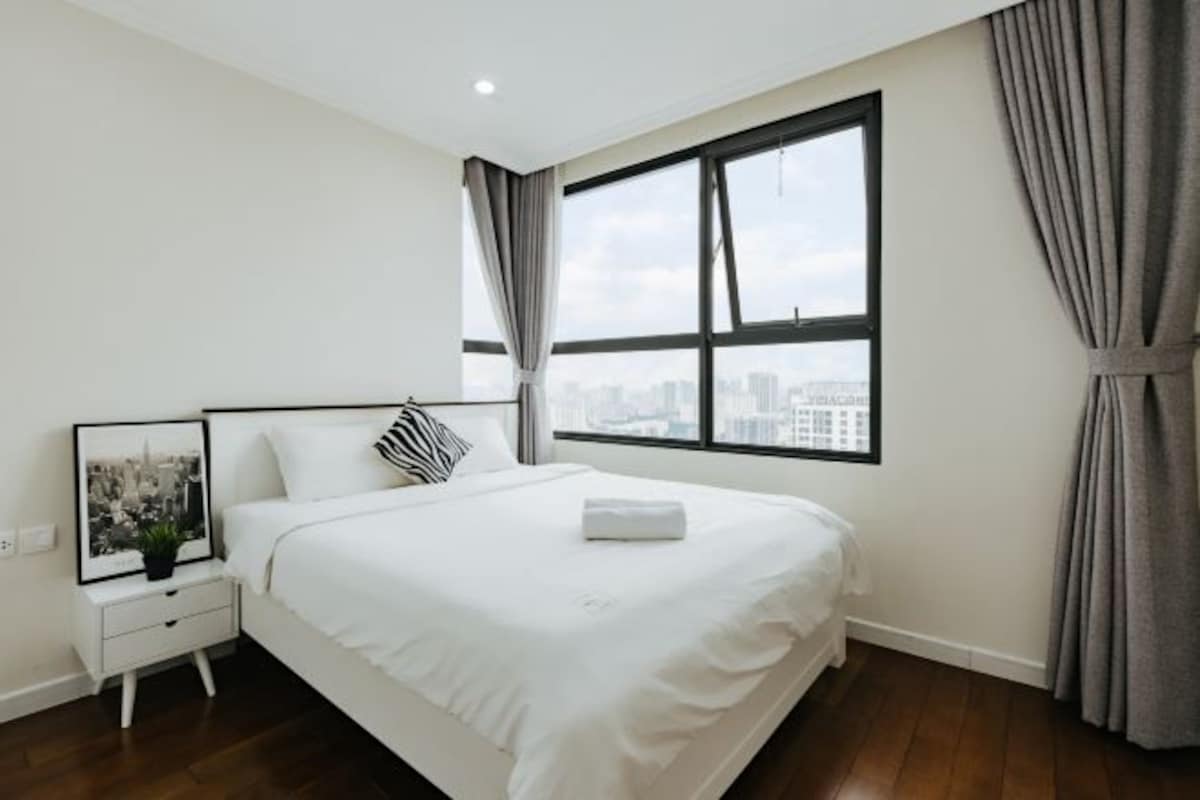 A modern bedroom with a large window, a neatly made bed, a bedside table with a framed picture, and a city view.