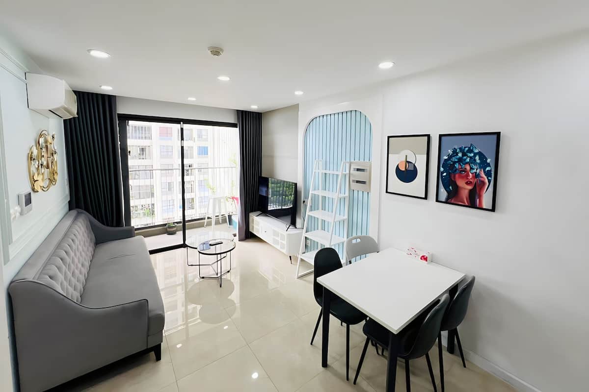 A modern living room with a grey sofa, a dining table with chairs, a TV, and a balcony with a view of buildings.