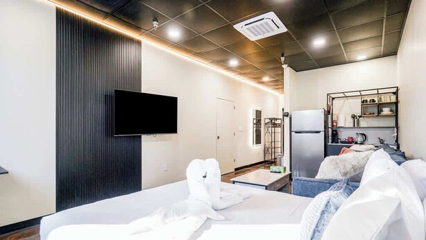 A modern hotel room with a large bed, a flat-screen TV mounted on the wall, a kitchenette with a refrigerator and microwave, and a small seating area with a sofa and cushions. - *New* Loft 3 min to Local hospitals Sleeps 4 (Pittsburgh)