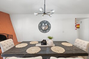 Un comedor con una mesa de madera oscura, sillas blancas tapizadas, una lámpara de araña negra, un espejo redondo y una pequeña planta en maceta.