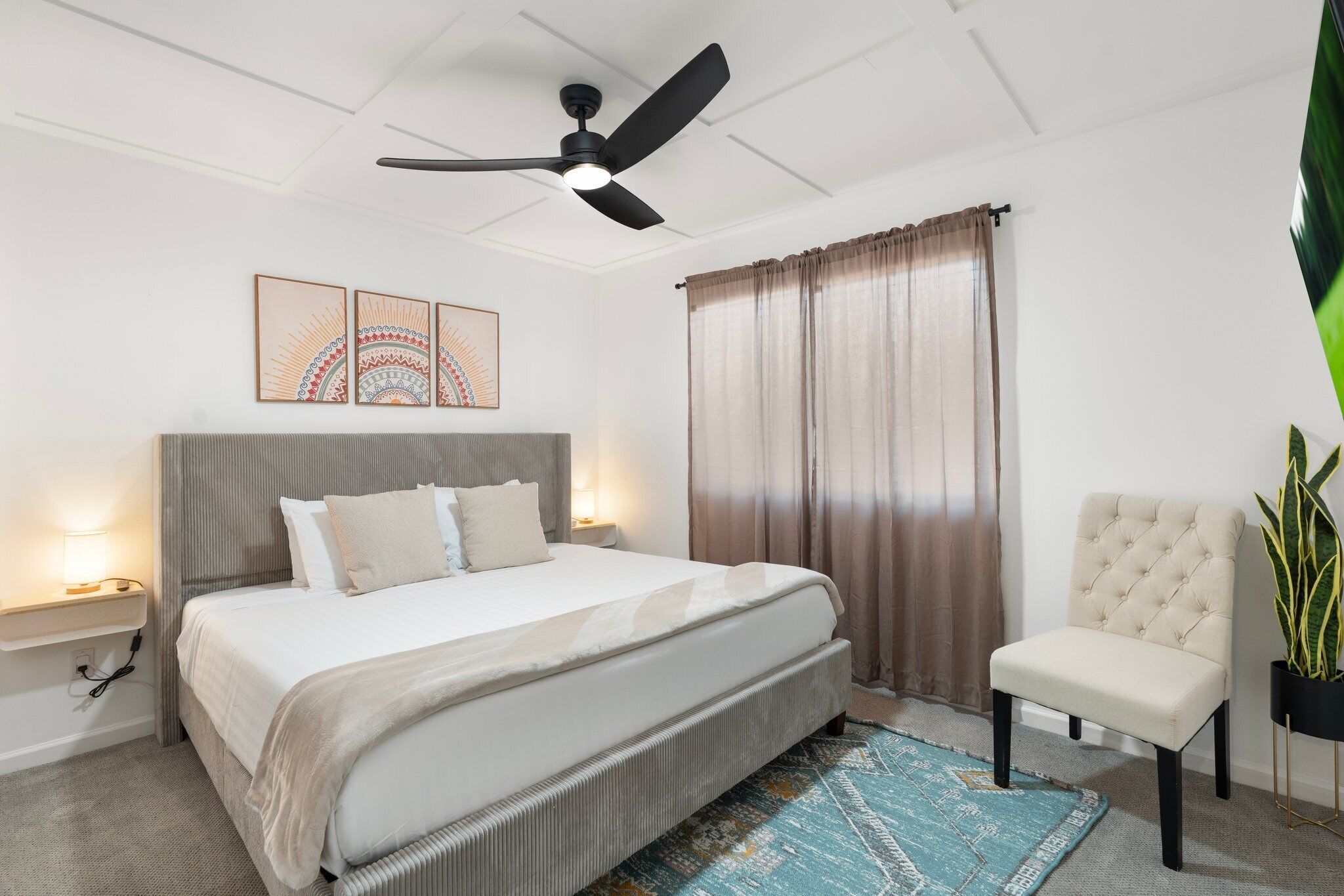 A bedroom with a bed, bedside lamps, a ceiling fan, a chair, and a plant.