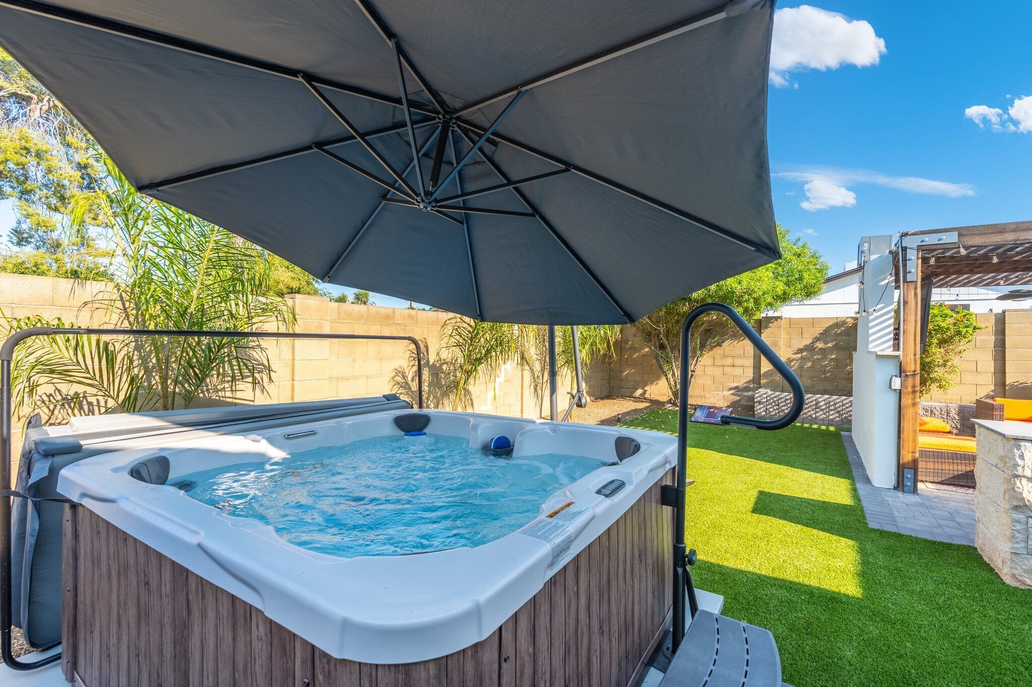 A hot tub under a large umbrella in a backyard with artificial grass and a brick wall.