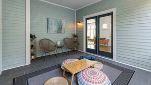 A covered patio area with a wooden table and chairs, a small shelf with plants, and a sliding glass door leading to an outdoor seating area. - Walk to Five Points | 2 Patios + Private Garage       |     Close to Dining and Bars, Board Games, 10 Mins to Broadway, 24 Hour Hospitality (Nashville)