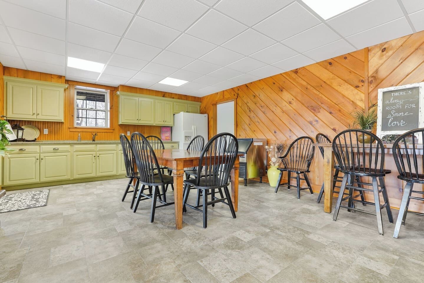 A dining area with a wooden table and chairs, a kitchen with green cabinets, and a chalkboard with a message.