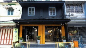 A two-story building with a black facade and yellow accents. There are two large windows on the upper floor and a door with glass panels. The entrance has a yellow awning and potted plants.