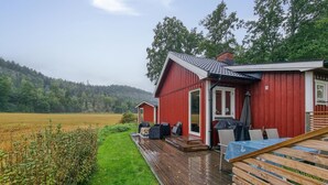 Une cabane rouge avec une terrasse en bois, un espace salon couvert et une vue sur un champ et une forêt en arrière-plan.
