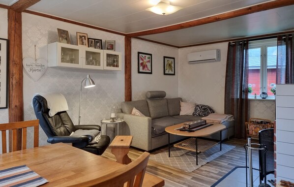 Un salon chaleureux avec un canapé, une table à manger et une chaise. On y trouve une lampe, une petite table d’appoint et une bibliothèque avec des photos encadrées.