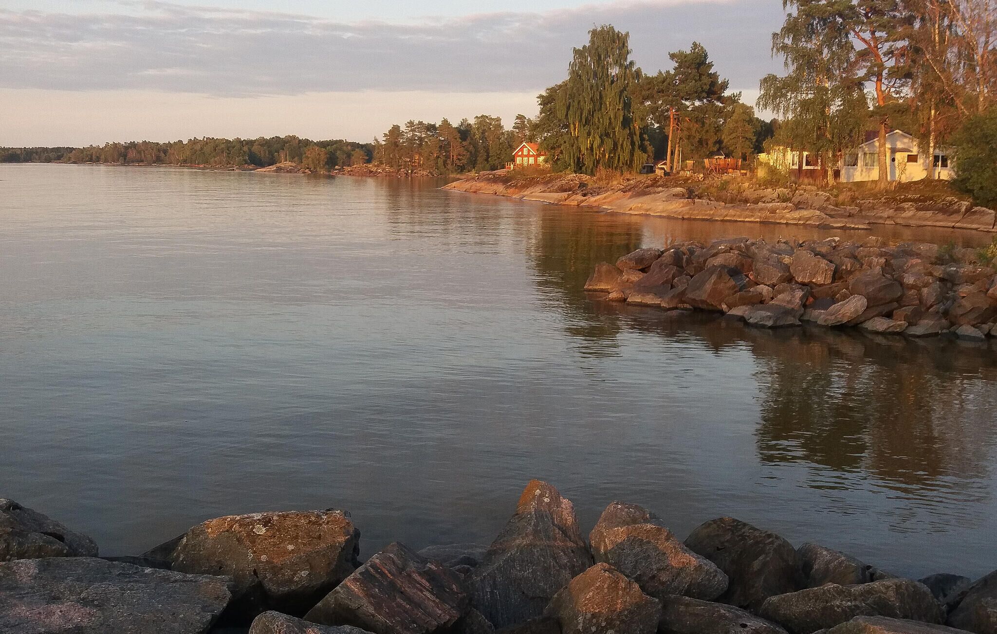Un paysage paisible au bord de l’eau, avec une côte rocheuse, une petite maison et des arbres en arrière-plan.