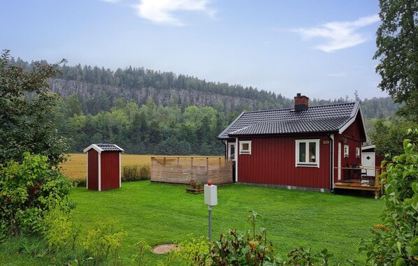 Une maison rouge avec un toit en tuiles, une terrasse en bois et un petit abri de jardin, le tout situé dans une cour verdoyante, avec une colline boisée en arrière-plan.