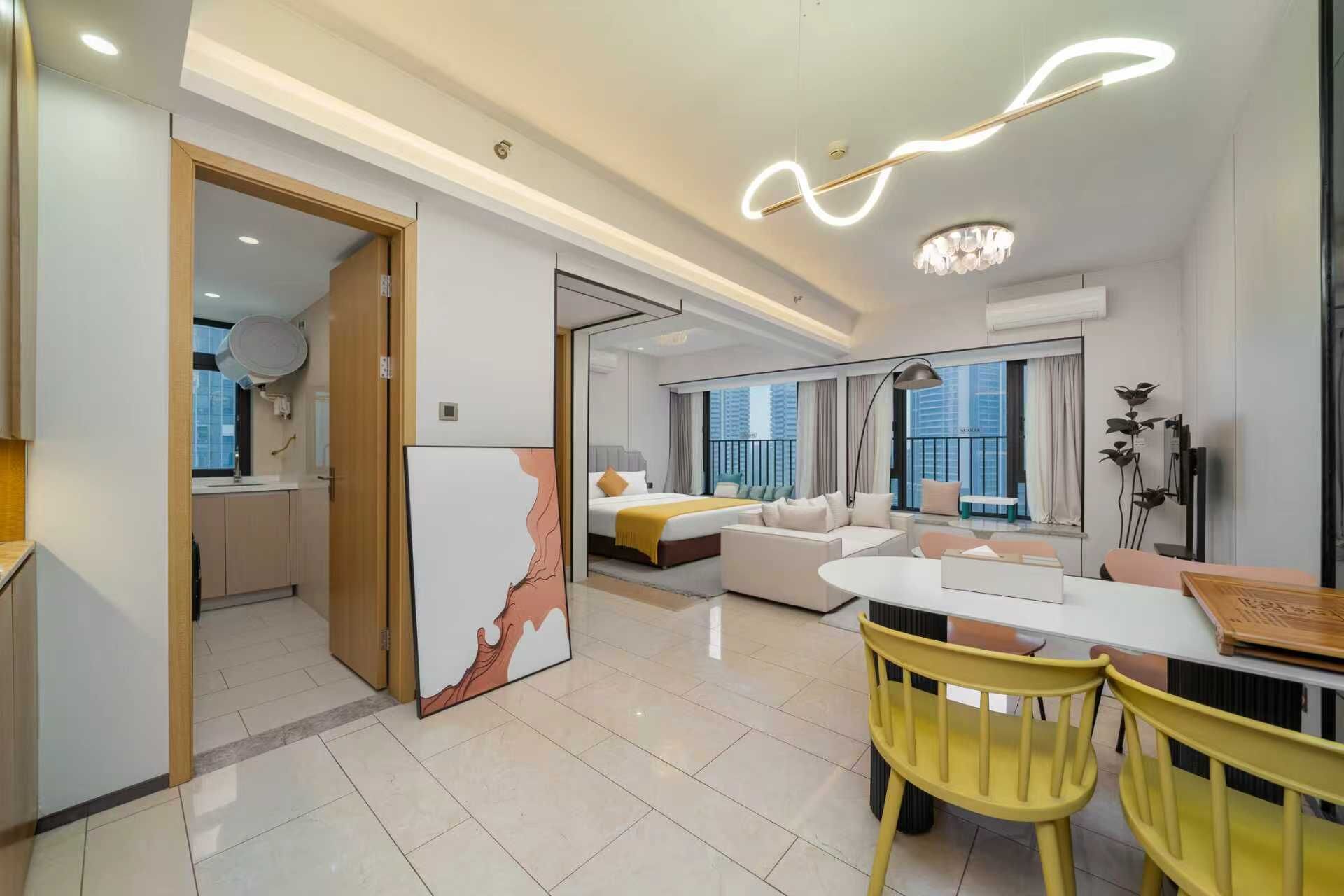 A modern hotel room with a large bed, a sofa, a dining area with yellow chairs, and a view of the city.