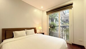 Een hotelkamer met een bed, een nachtkastje, een lamp, een raam met uitzicht en een balkon met planten.