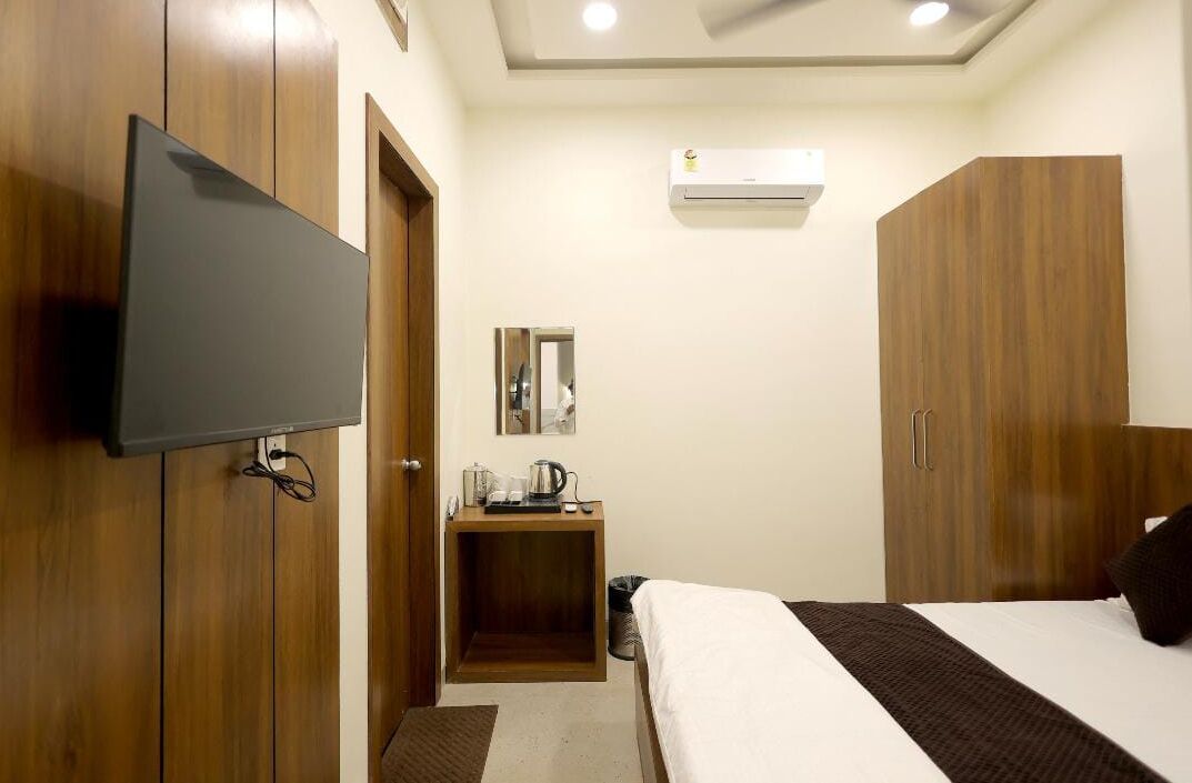 A hotel room with a flat-screen TV mounted on the wall, a wooden wardrobe, a bedside table with a kettle, and a bed with a white and brown striped bedspread.