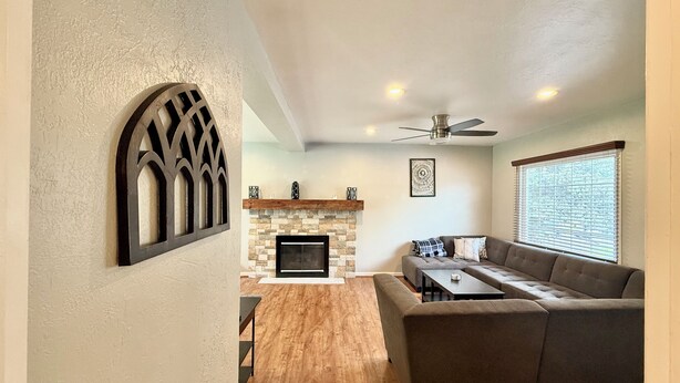 A living room with a fireplace, a ceiling fan, and a large window with blinds.