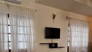 Una sala de estar con sofás color beige, una mesa de centro negra, un televisor de pantalla plana montado en la pared y cortinas translúcidas en las ventanas.