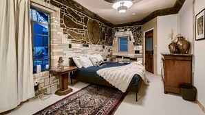 A bedroom with a stone-patterned wall, a bed with a blue bedspread, a wooden nightstand, a large vase, and a ceiling fan. - Historic Artist Home-Walk to SXSW+6th St+UT (Austin)