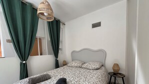 Une chambre à coucher avec un lit, des rideaux verts, un luminaire en osier, une petite table de chevet et une grille d’aération fixée au mur.